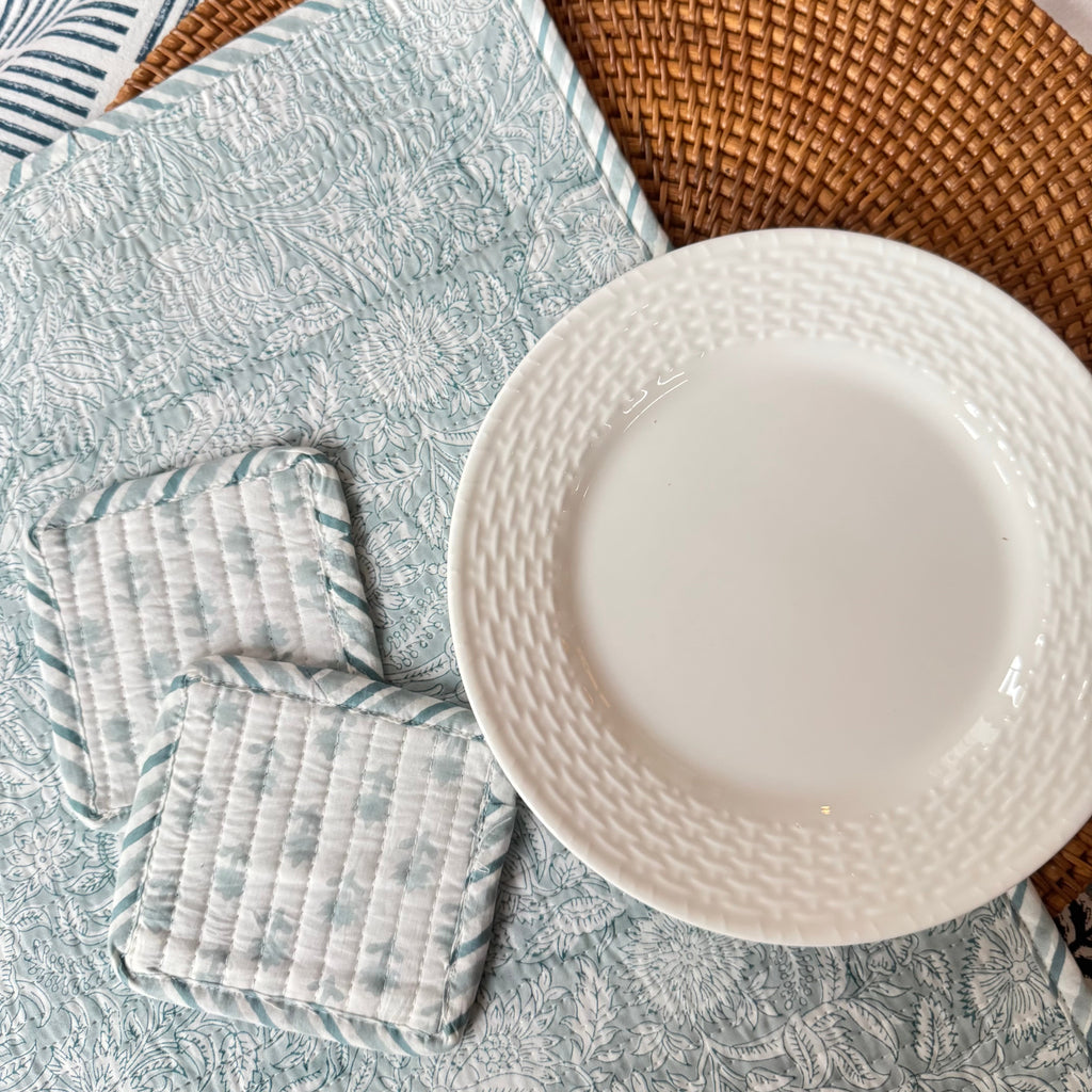 White plate on top of wicker and floral patterned placemats next tow two floral patterned coasters, on top of table cloth with floral and geometric design.