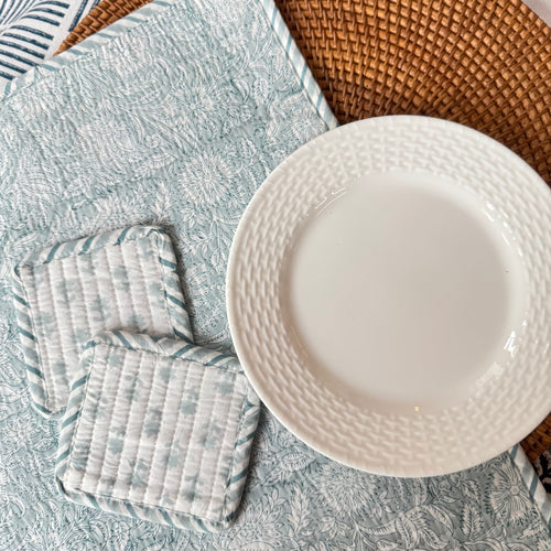 White plate on top of wicker and floral patterned placemats next tow two floral patterned coasters, on top of table cloth with floral and geometric design.