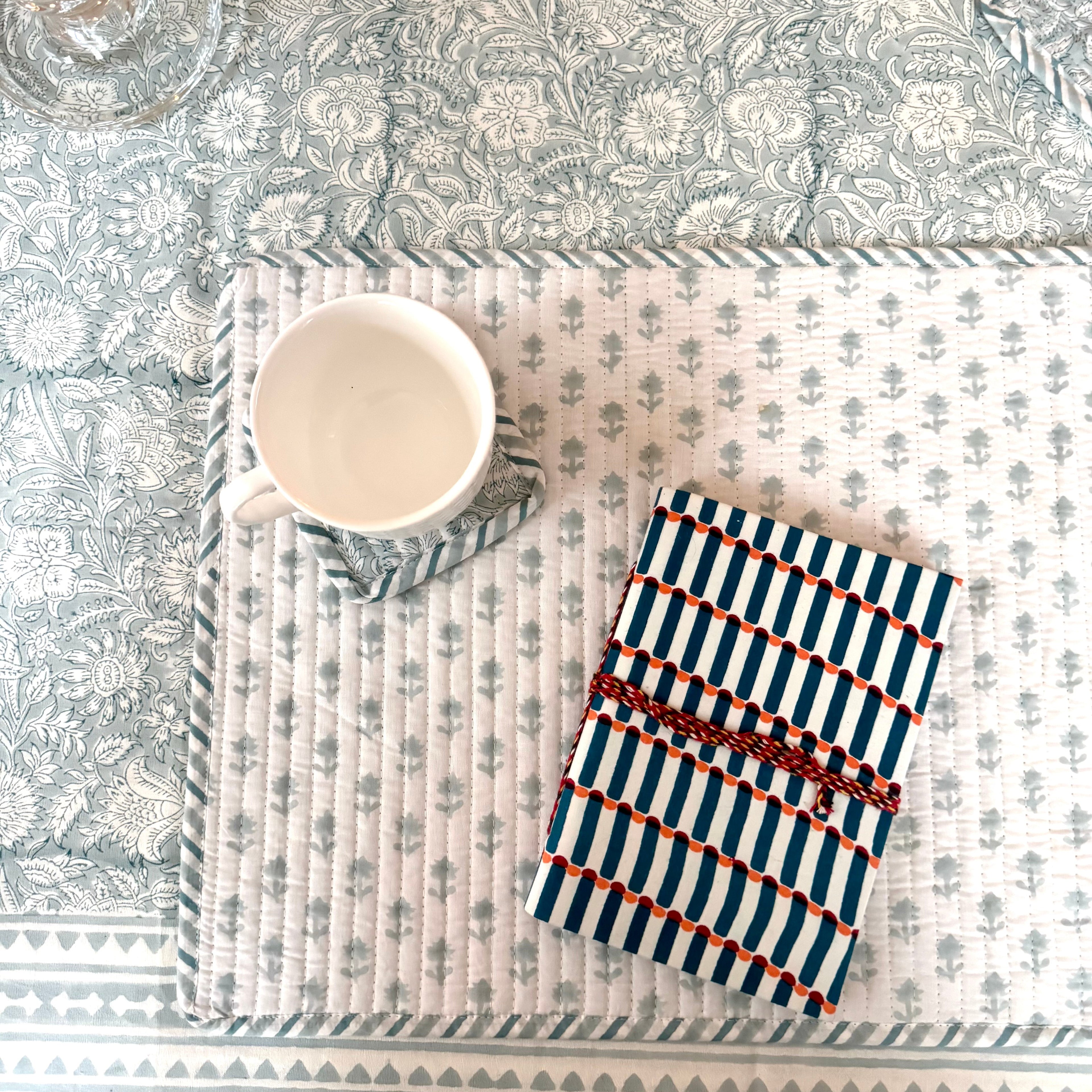 Geometric patterned notebook on floral placemat with white mug on top of floral coaster, on floral patterned table cloth.