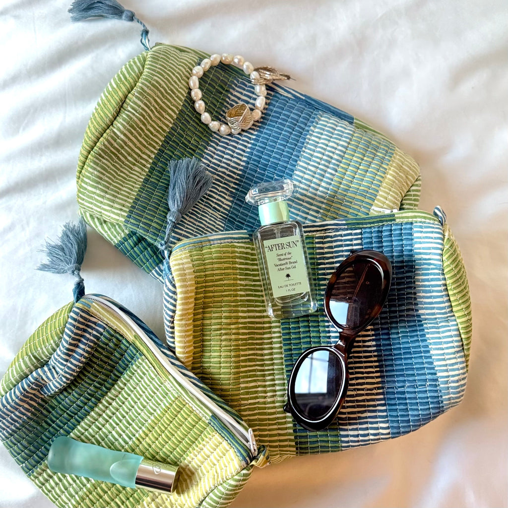 Three striped cosmetic bags on white bed spread with various perfumes, brown sunglasses, pearl bracelet, gold ring, and gold earrings.