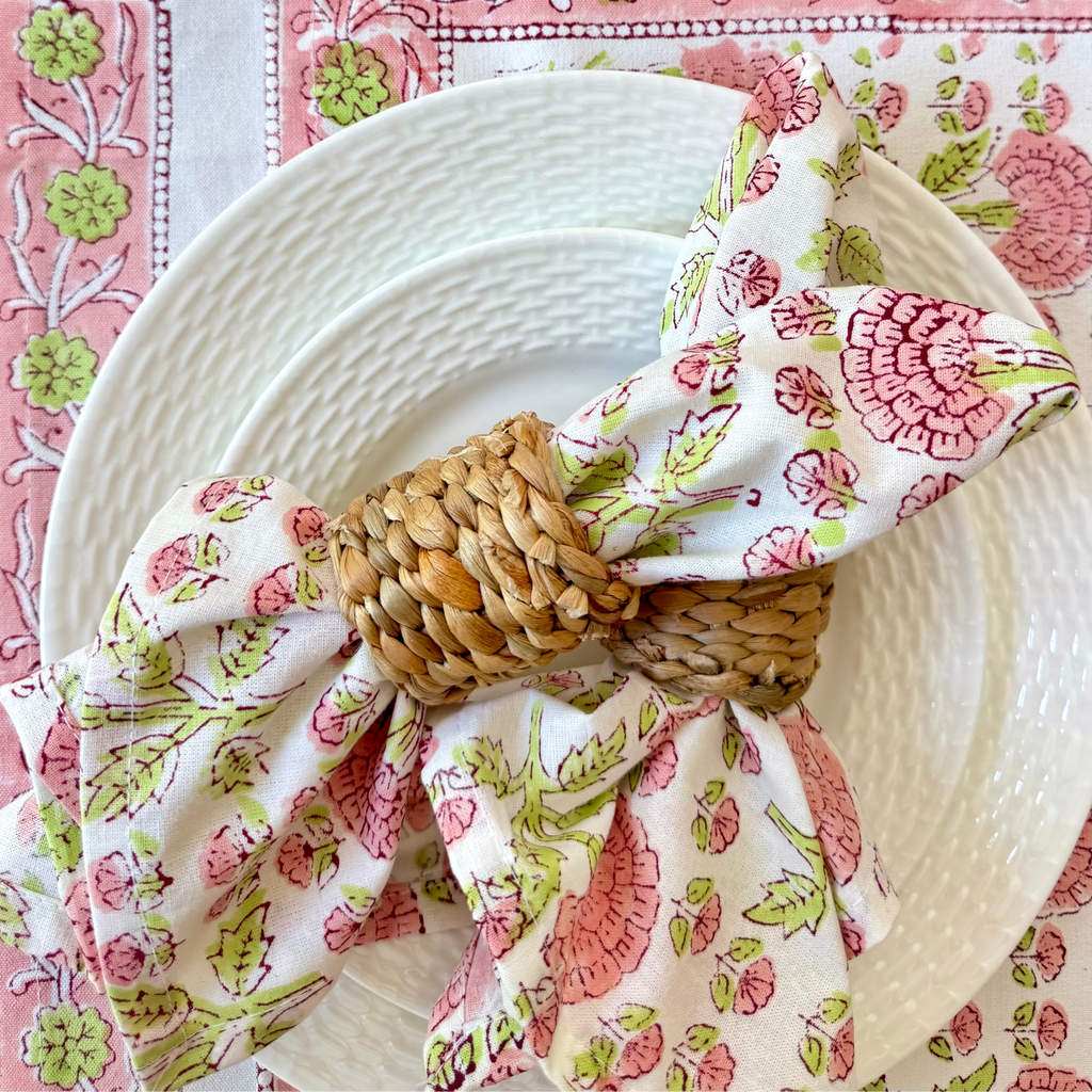 Decorative table setting with floral napkin and woven napkin ring on a pink and green patterned tablecloth.