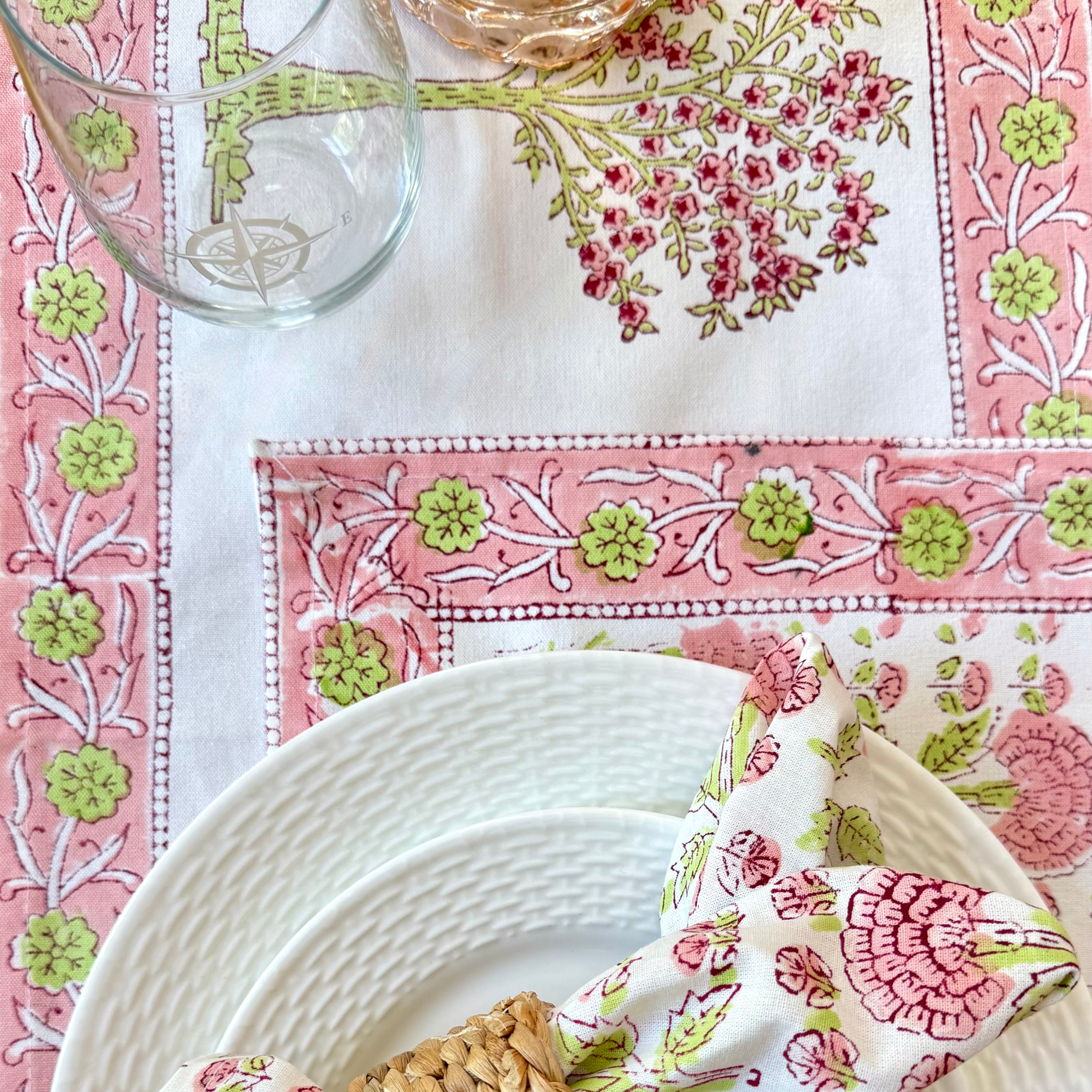Decorative table setting with floral napkin and woven napkin ring and glass on a pink and green patterned tablecloth.