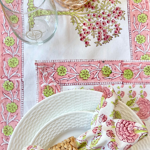 Decorative table setting with floral napkin and woven napkin ring and glass on a pink and green patterned tablecloth.