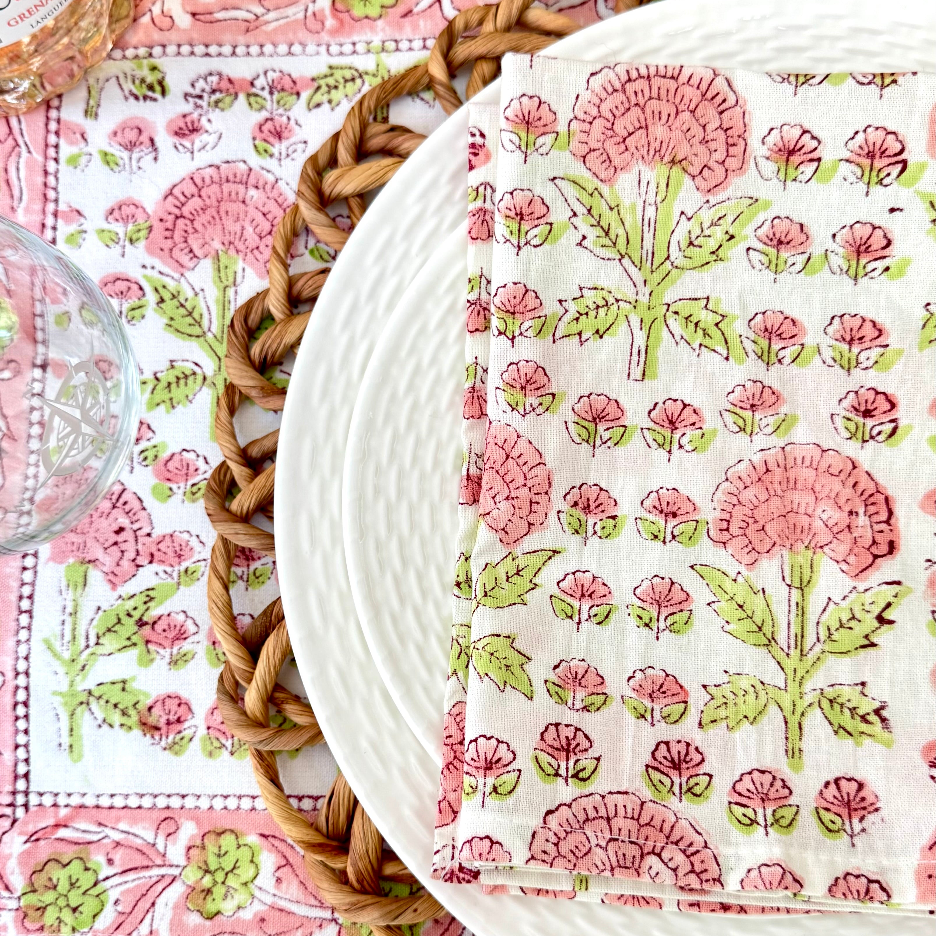 Floral-patterned napkin on a matching placemat with a woven placemat underneath with two white plates a glass and a bottle of wine.