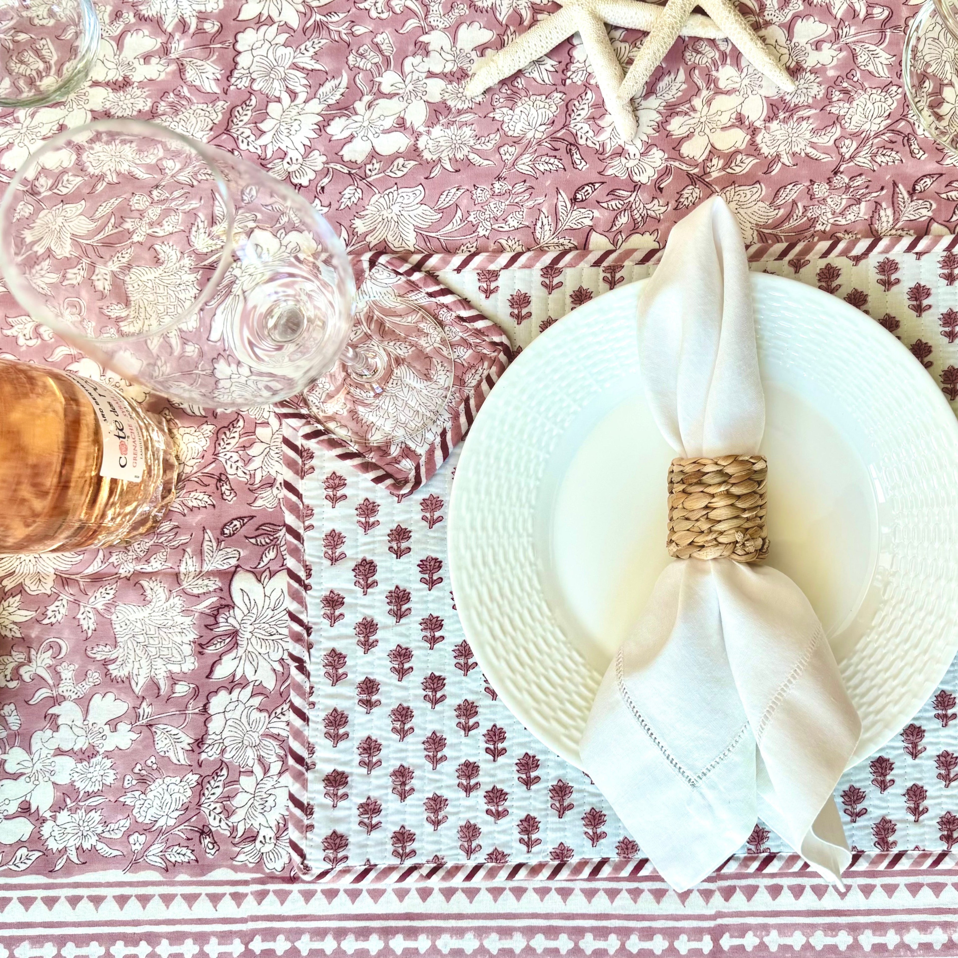 Table setting with a white plate, folded napkin, and decorative ring on a pink floral tablecloth.