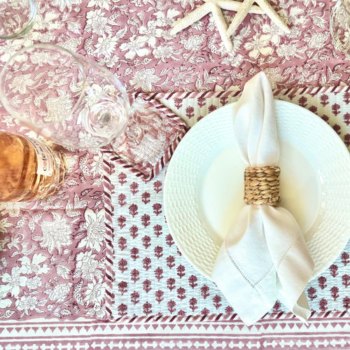 Table setting with a white plate, folded napkin, and decorative ring on a pink floral tablecloth.