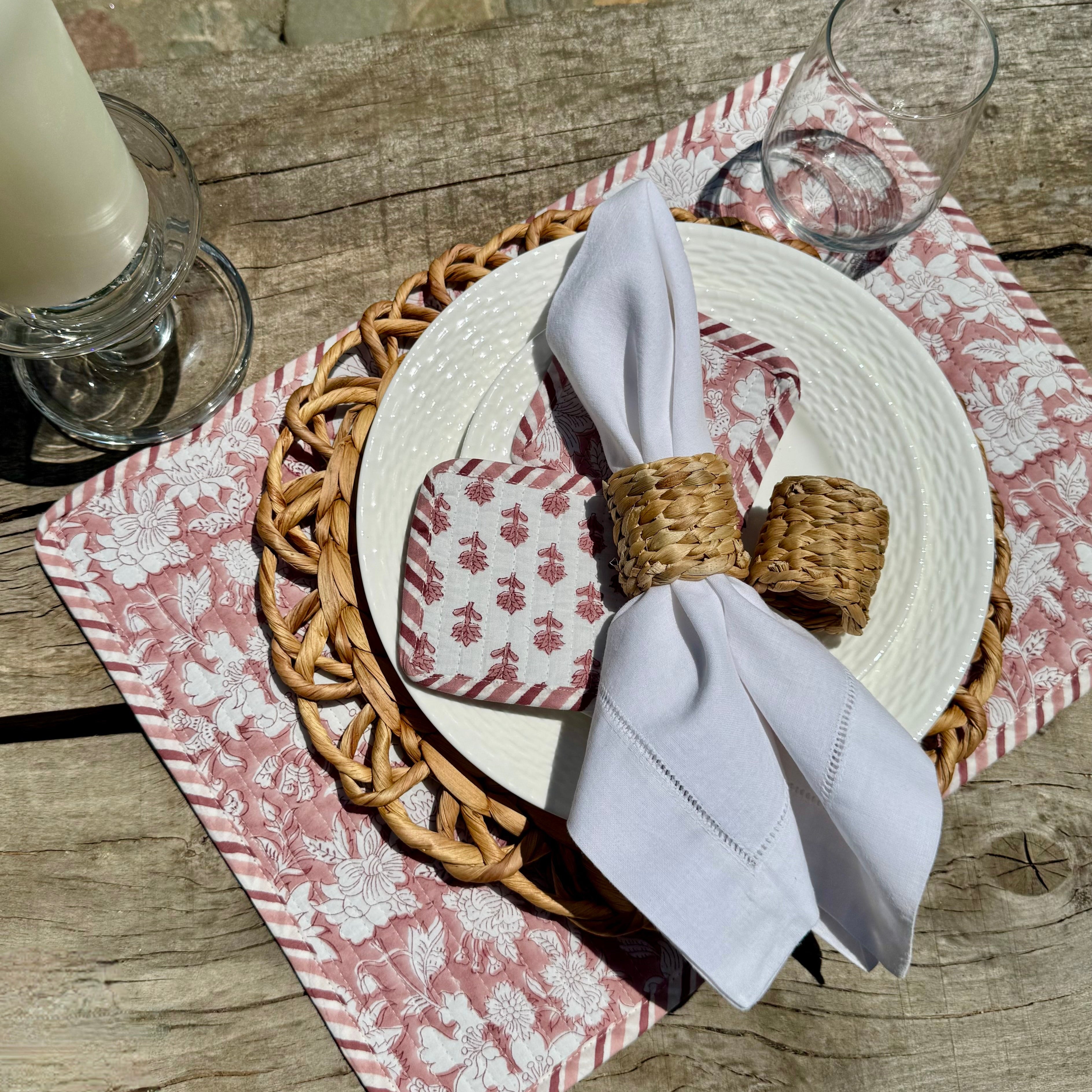 Table setting with plates, napkins, coasters, woven napkin rings, candle, wine glass, and a woven charger on a wooden table.