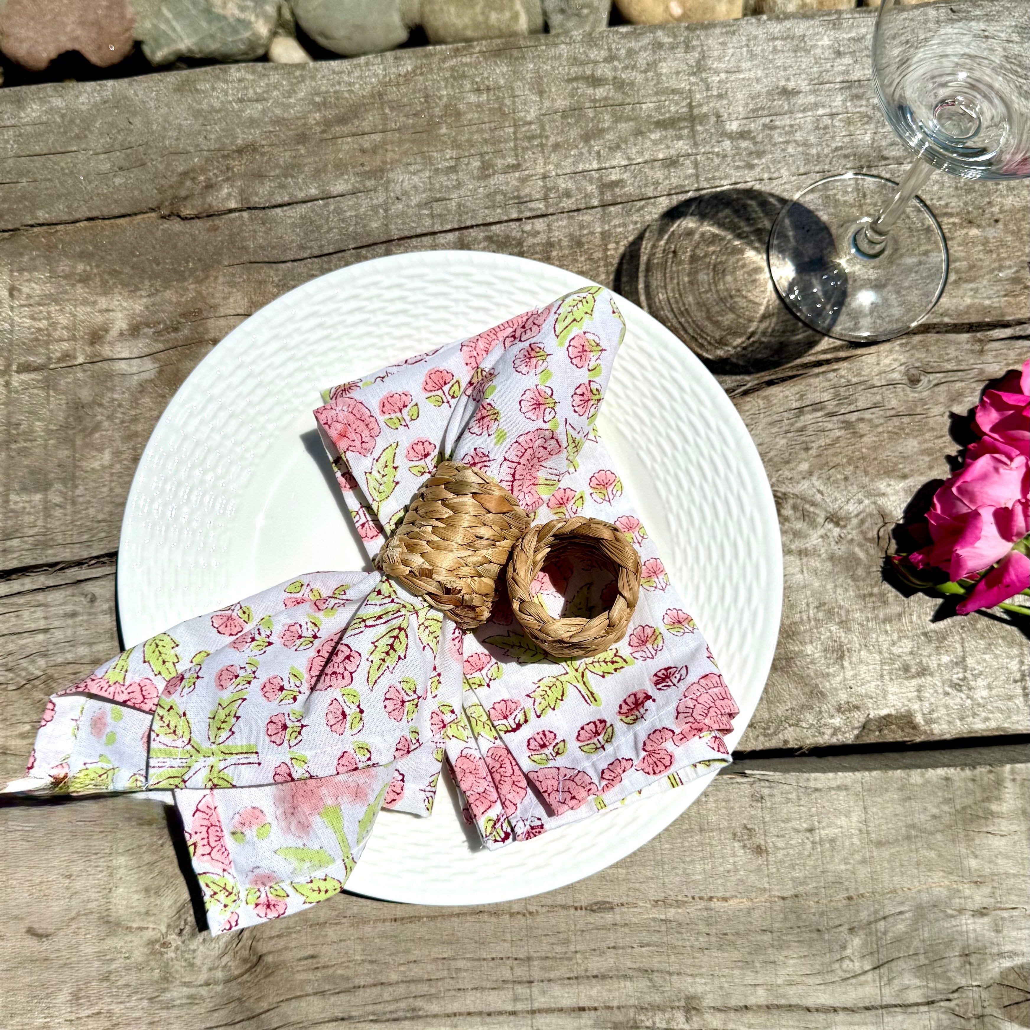 Floral napkins on a white plate with woven napkin rings a glass and flowers on a wooden surface next to a stone wall