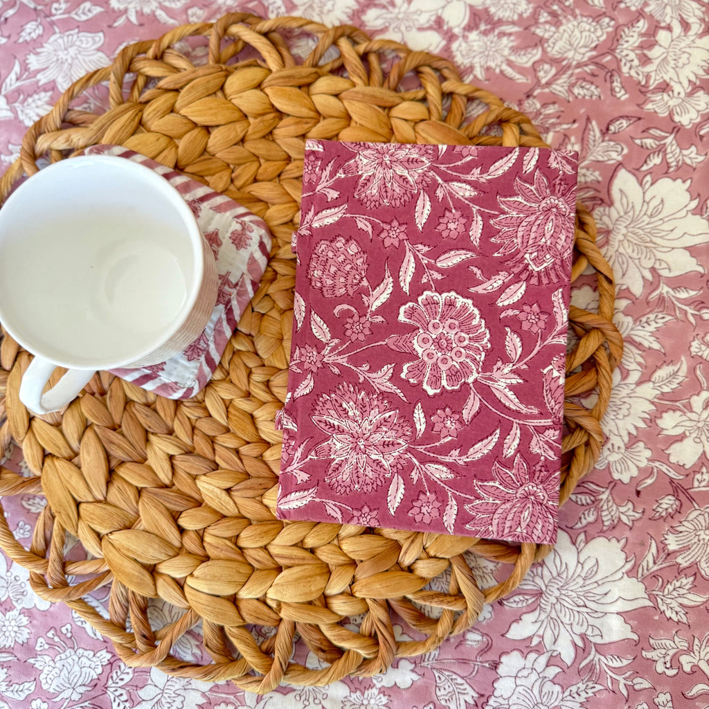 Woven placemat with a floral-patterned notebook, and white mug on a pink floral coaster on a pink floral tablecloth.