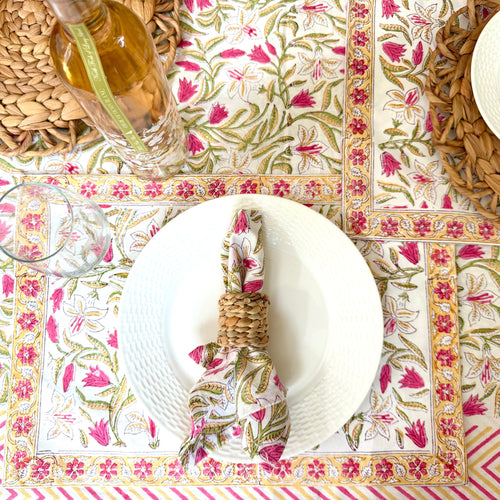 A styled tabletop setting featuring a floral tablecloth and matching placemats in pink, green, and gold, with white plates, woven napkins, and natural woven placemats.