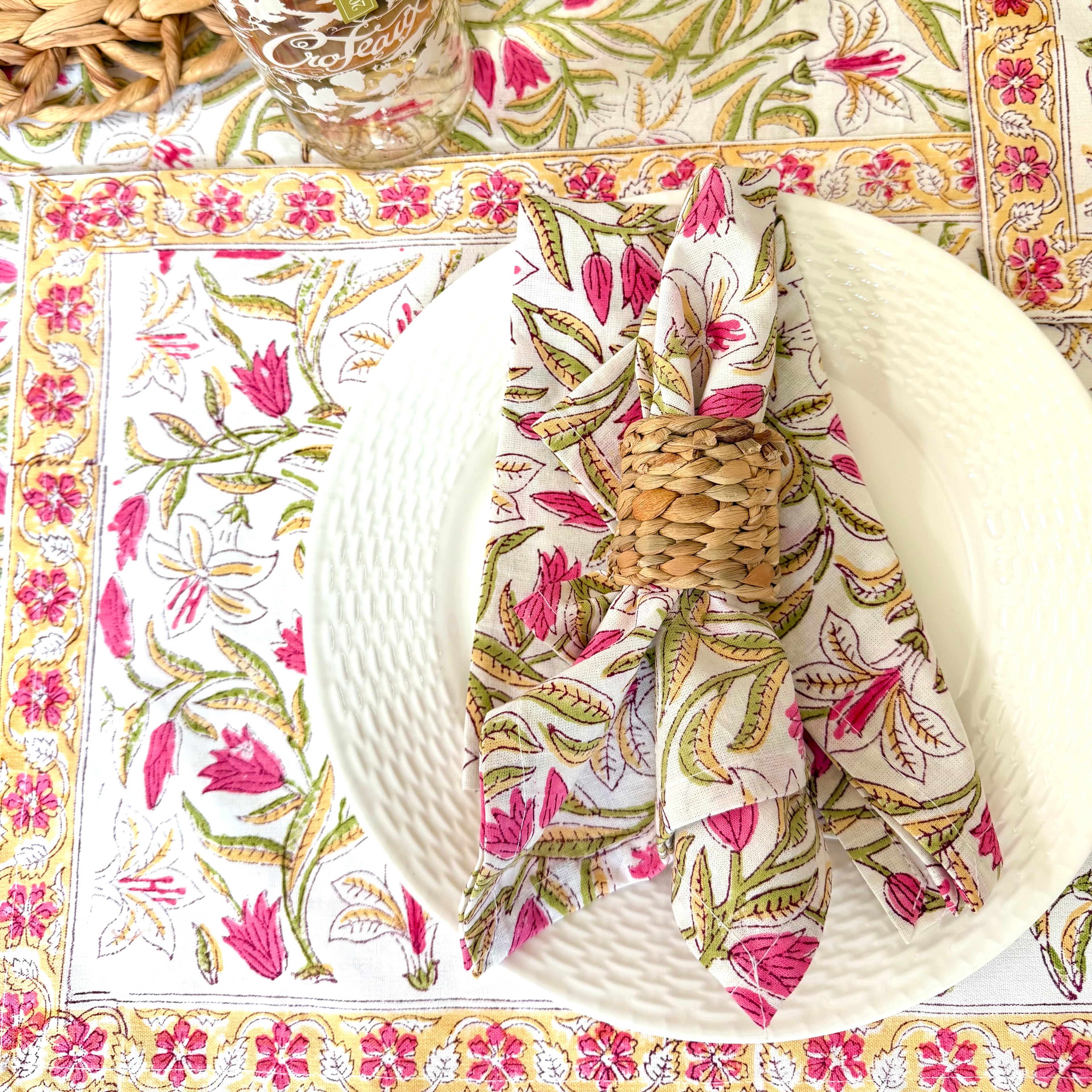 A styled table setting featuring a white textured plate, a folded napkin with a pink and green floral block print, and matching printed placemats and a glass on a woven placemat.