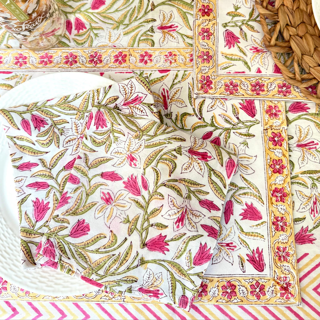 A styled flat-lay of a matching table setting featuring a white tablecloth and napkins with a vibrant pink and green botanical block print, bordered by gold and pink patterns. The setting includes woven natural fiber placemats, a glass jar, and a white plate.