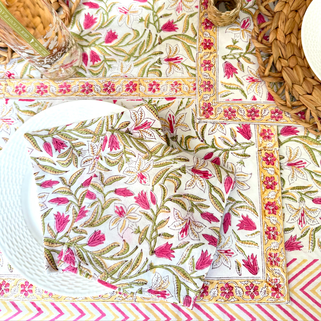 A styled table setting featuring a tablecloth and matching napkins with a vibrant pink and green floral block print, bordered by a pink and yellow chevron pattern. The setting includes woven straw placemats, white plates, and a glass bottle on a neutral tabletop.