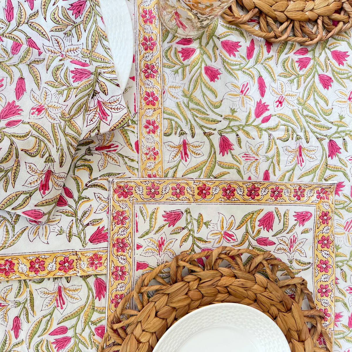 A styled flat-lay of a matching table setting featuring a white tablecloth and napkins with a vibrant pink and green floral block-print pattern, accented with gold borders and woven natural placemats and a white plate.