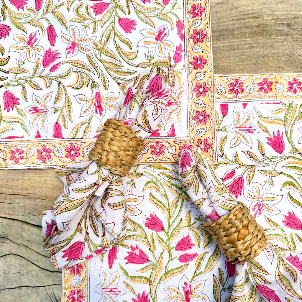 A set of white table linens with a vibrant pink and gold floral block print, styled on a rustic wooden table with two matching napkins in woven napkin rings.