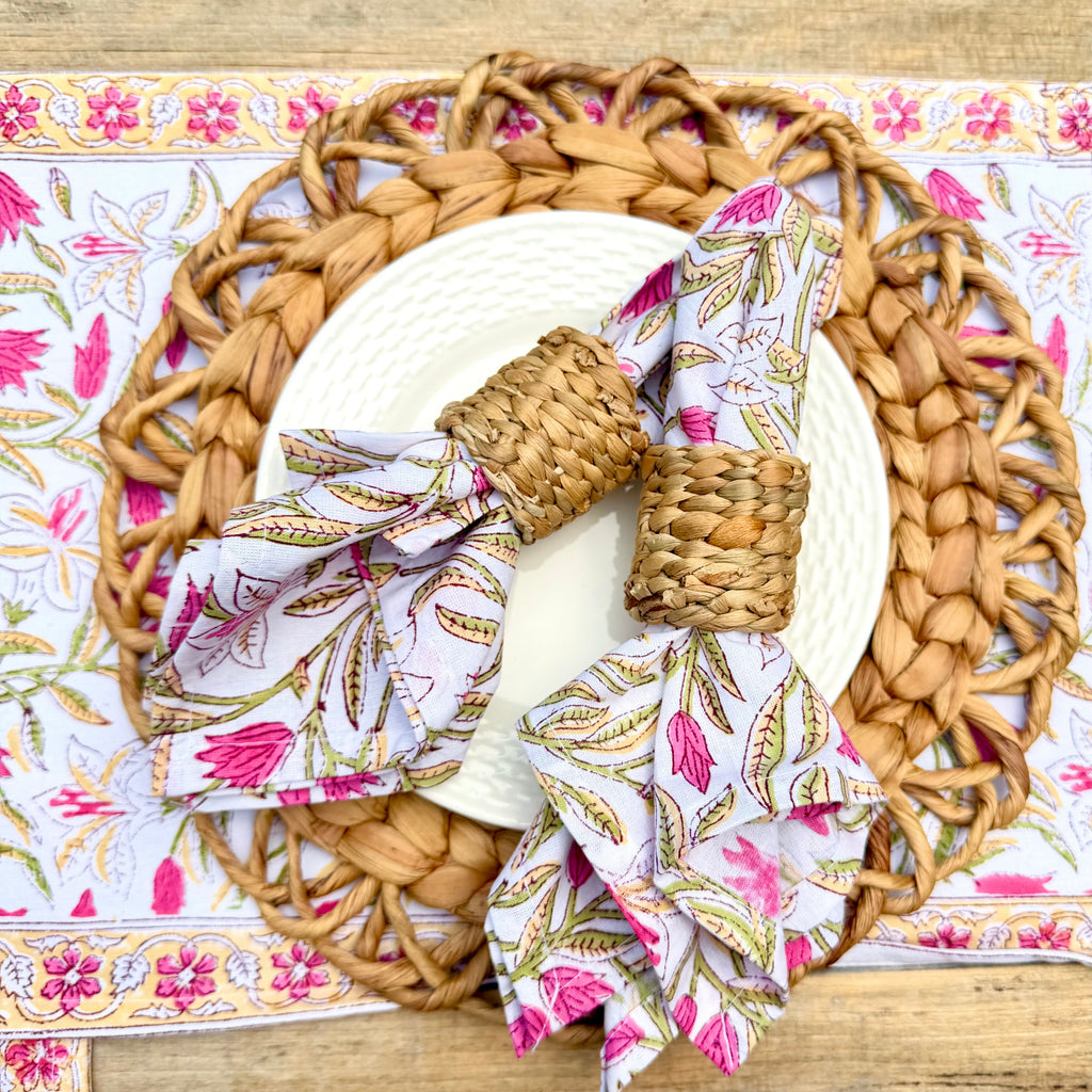A styled table setting featuring a round woven straw placemat, a white textured plate, a matching woven napkin ring, and matching patterned napkins with pink, green, and gold botanical prints, all arranged on a rustic wooden table.