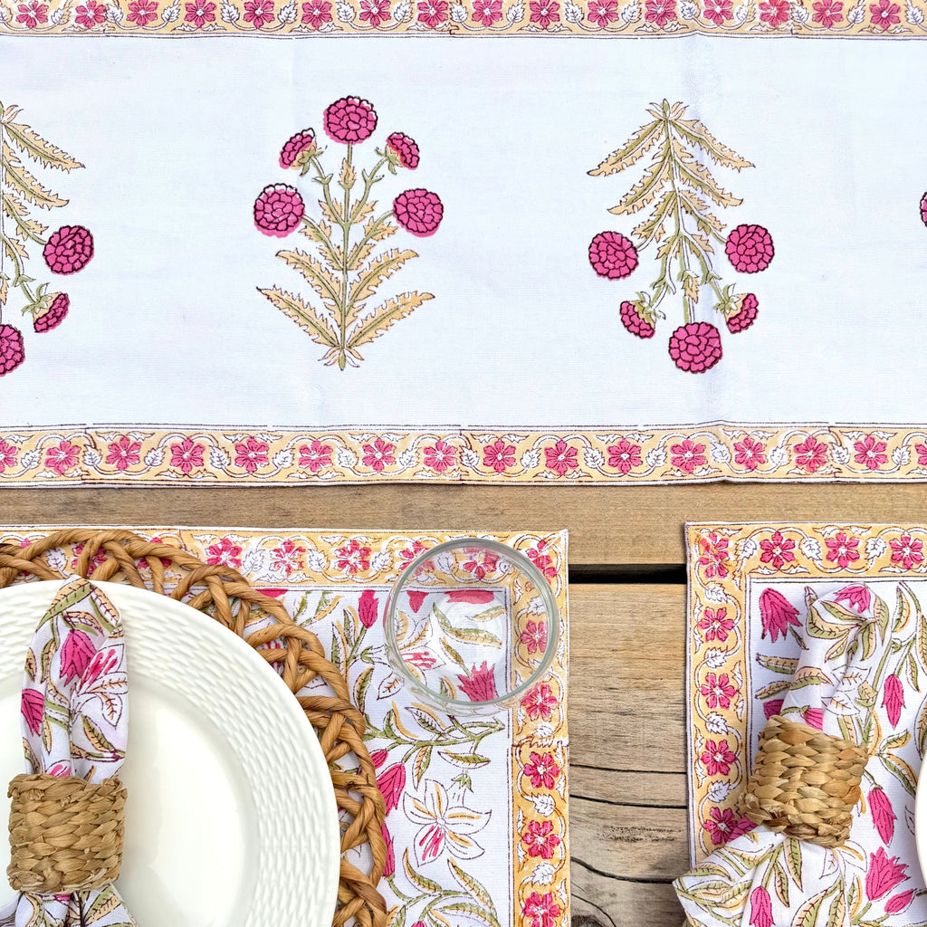 A styled table setting featuring a white table runner and matching placemats with pink and gold floral block-print patterns, set on a rustic wooden table with woven napkins and plates.