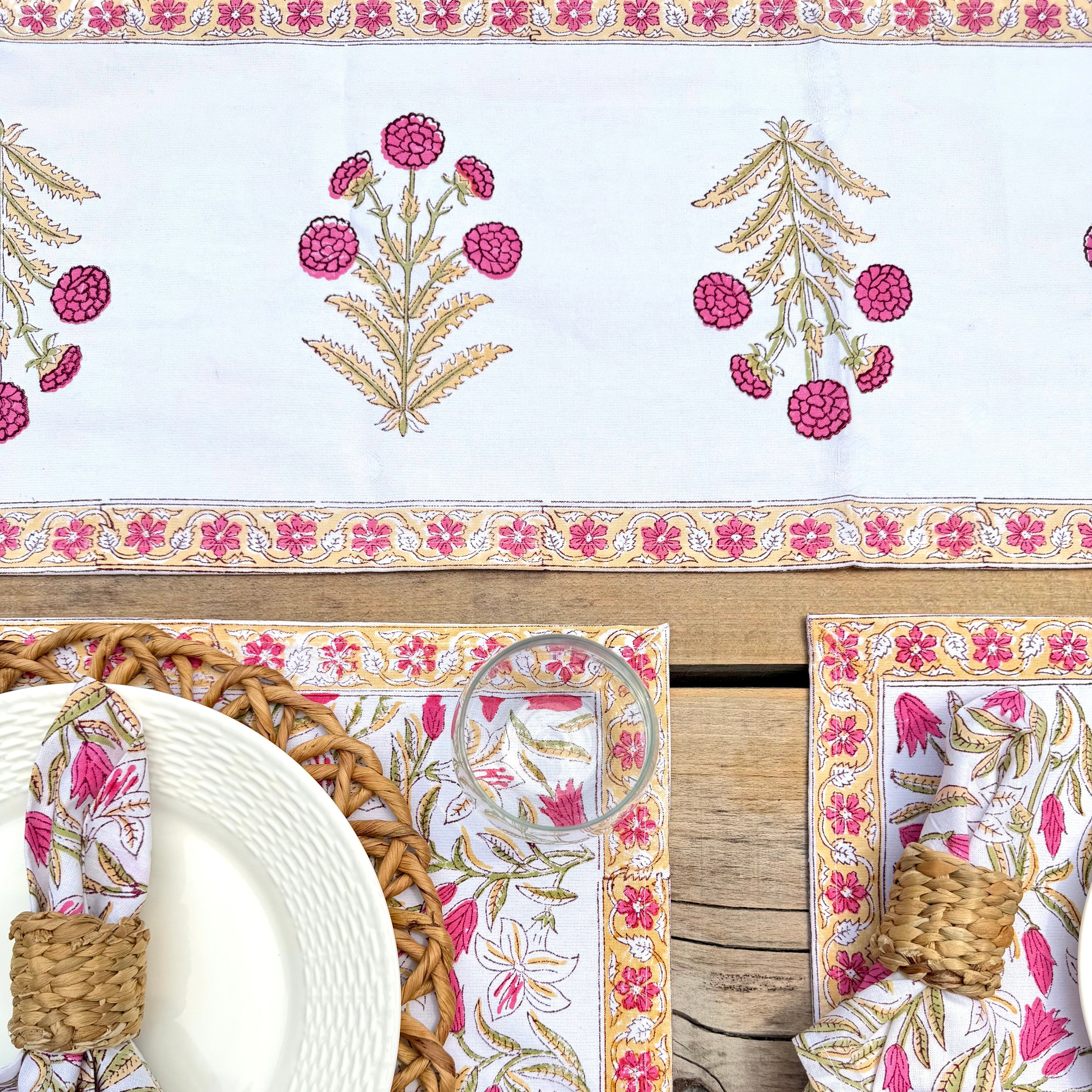 A styled table setting featuring a white table runner and matching placemats with pink and gold floral block-print patterns, set on a rustic wooden table with woven napkins and plates.