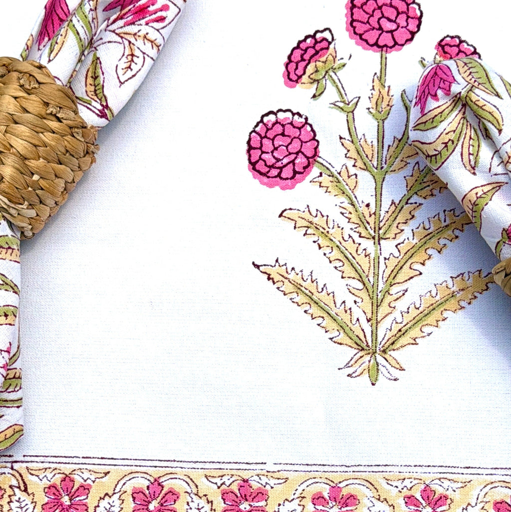 A close-up of a white table runner or tablecloth with a pink and gold floral border design, styled with woven placemats, a white plate, and floral napkins.