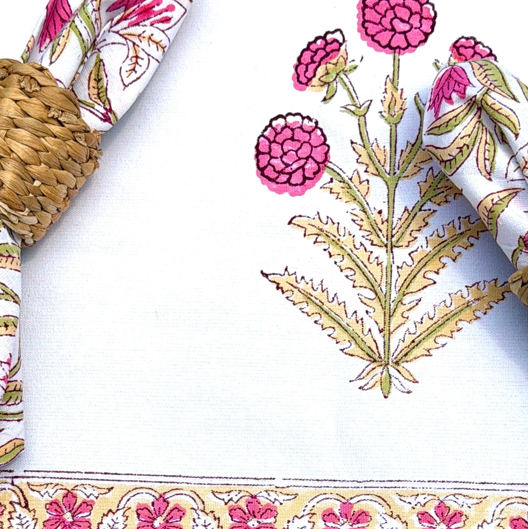 A close-up of a white table runner or tablecloth with a pink and gold floral border design, styled with woven placemats, a white plate, and floral napkins.