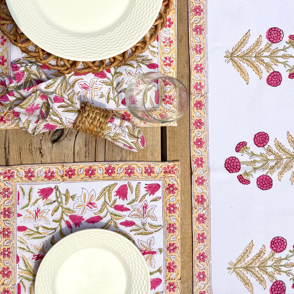 A styled flat-lay of a table setting featuring a white table runner and matching placemats with pink floral prints and gold botanical borders, alongside white textured plates, a glass, and woven napkins on a wooden table.