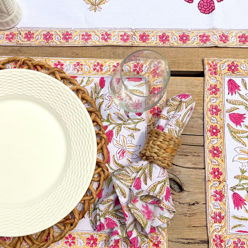 A styled flat-lay of a table setting featuring a white table runner and matching placemats with pink floral and green botanical patterns, alongside a white plate, woven placemat, glass, and a patterned napkin on a rustic wooden table.