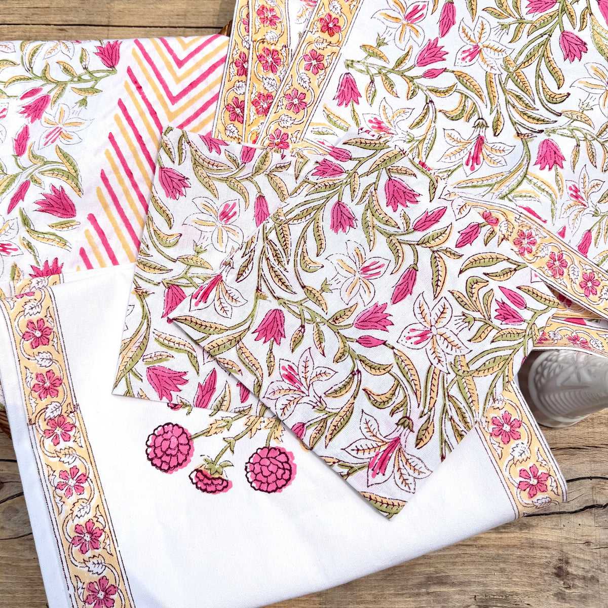 A folded white fabric garment or textile laid on a rustic wooden surface, featuring pink floral and leaf block-print patterns, gold outlines, striped accents, and embroidered pomegranate motifs near the border.