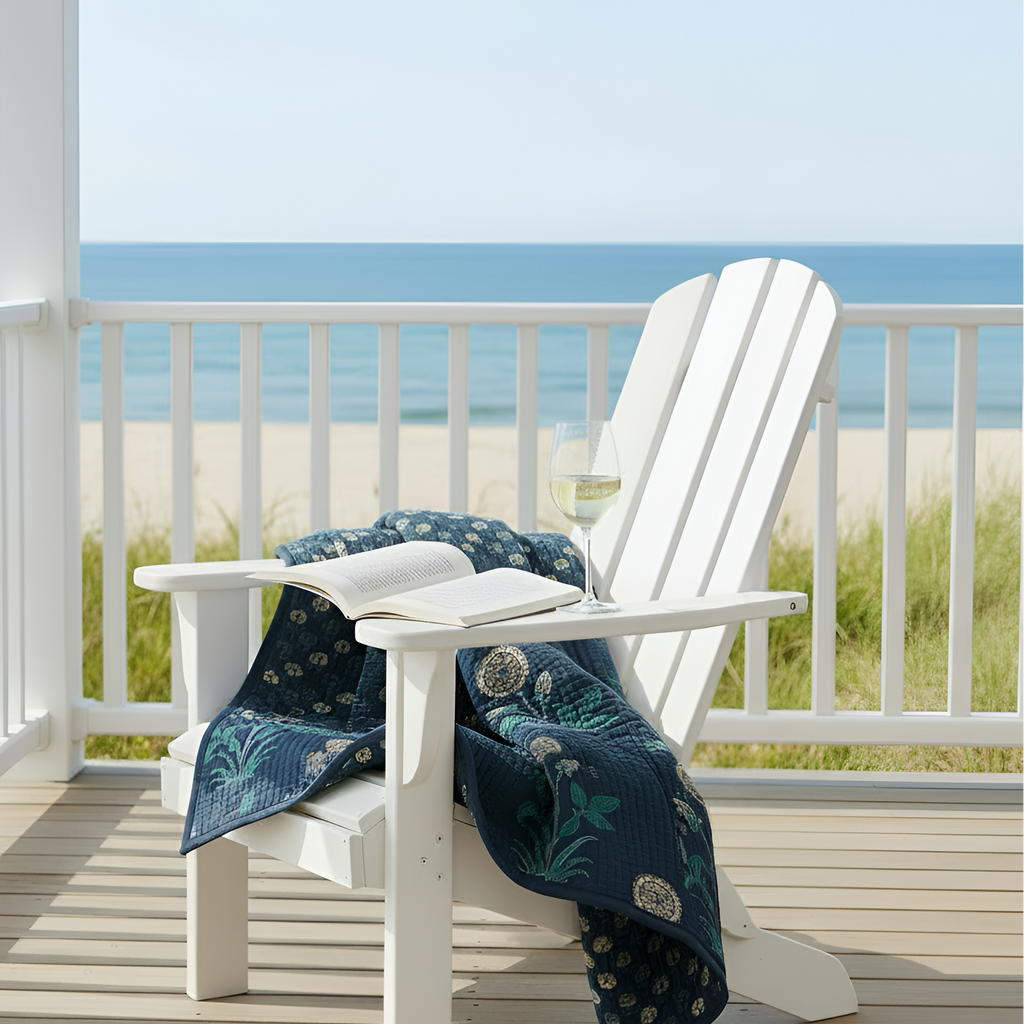 White Adirondack chair with a blue patterned blanket and an open book on a balcony with a beach view.