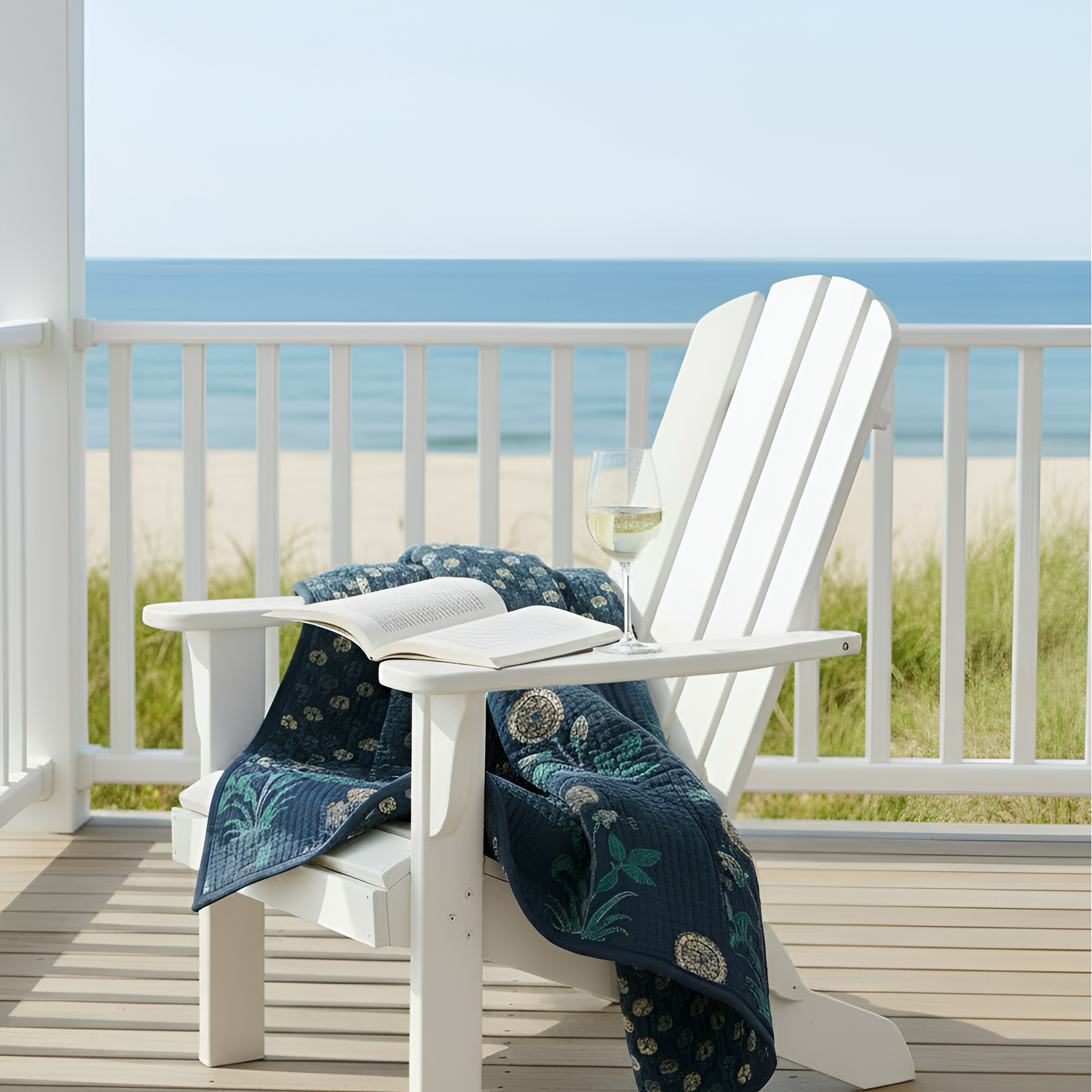 White Adirondack chair with a blue patterned blanket and an open book on a balcony with a beach view.