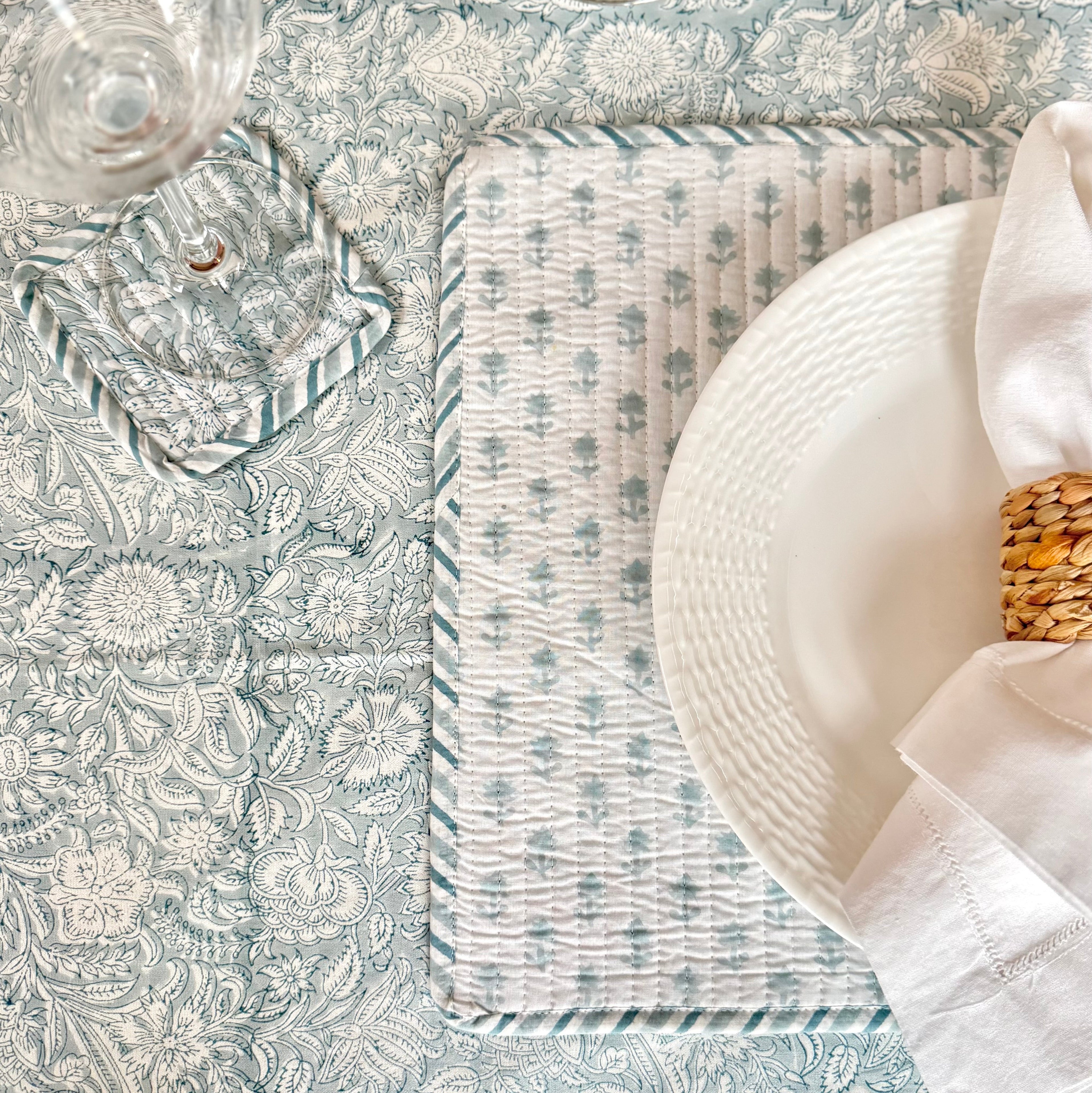 White napkin with sisal napkin ring on white plate on top of floral placemat, on floral patterned table cloth with wine glass on top of floral patterned coaster.