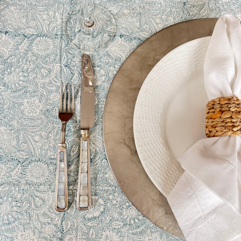 Dining table setting with white plates, silverware, white napkin, grey placemat, and wine glass on a patterned tablecloth.