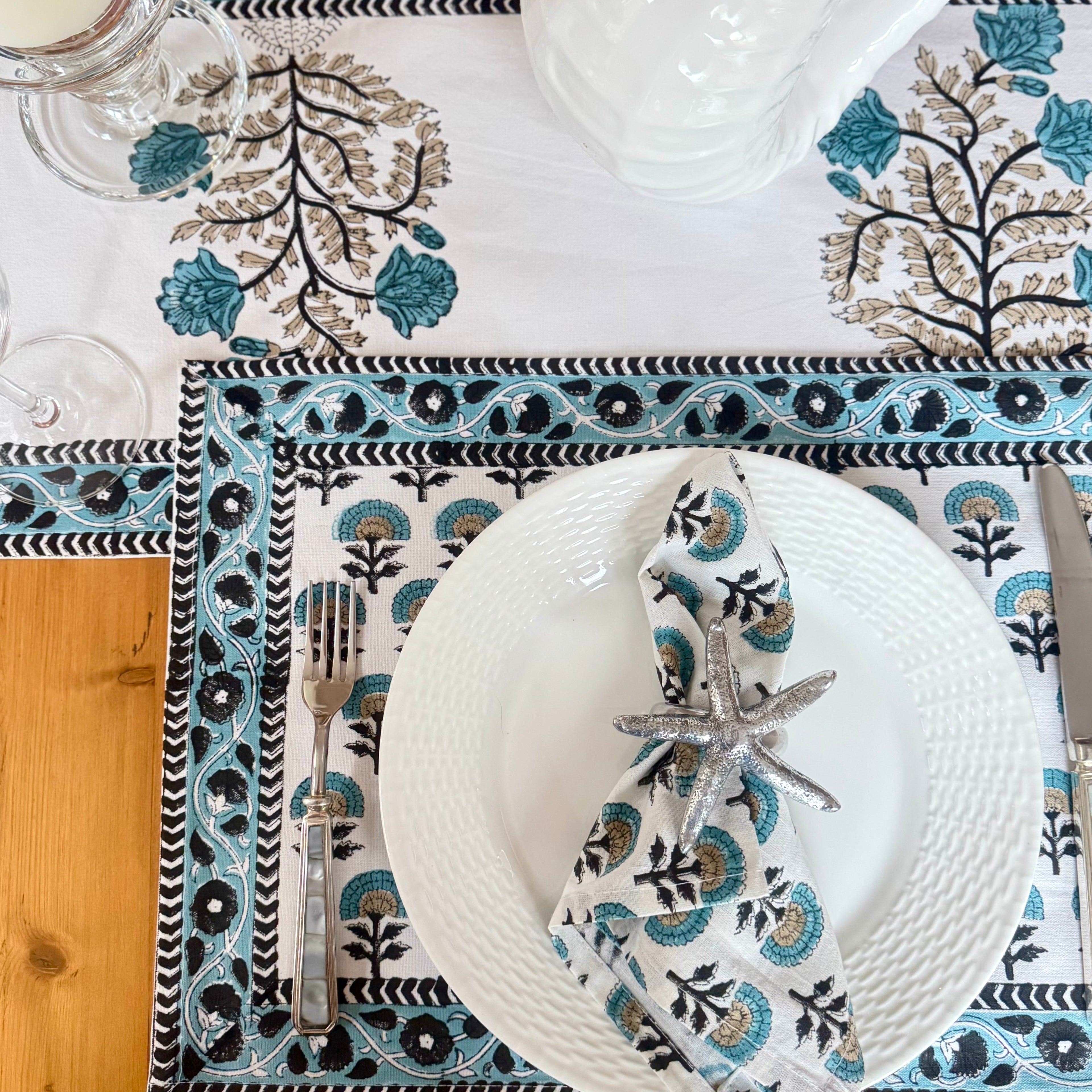 Table setting with floral placemats, white plates, and silver cutlery on a wooden table.